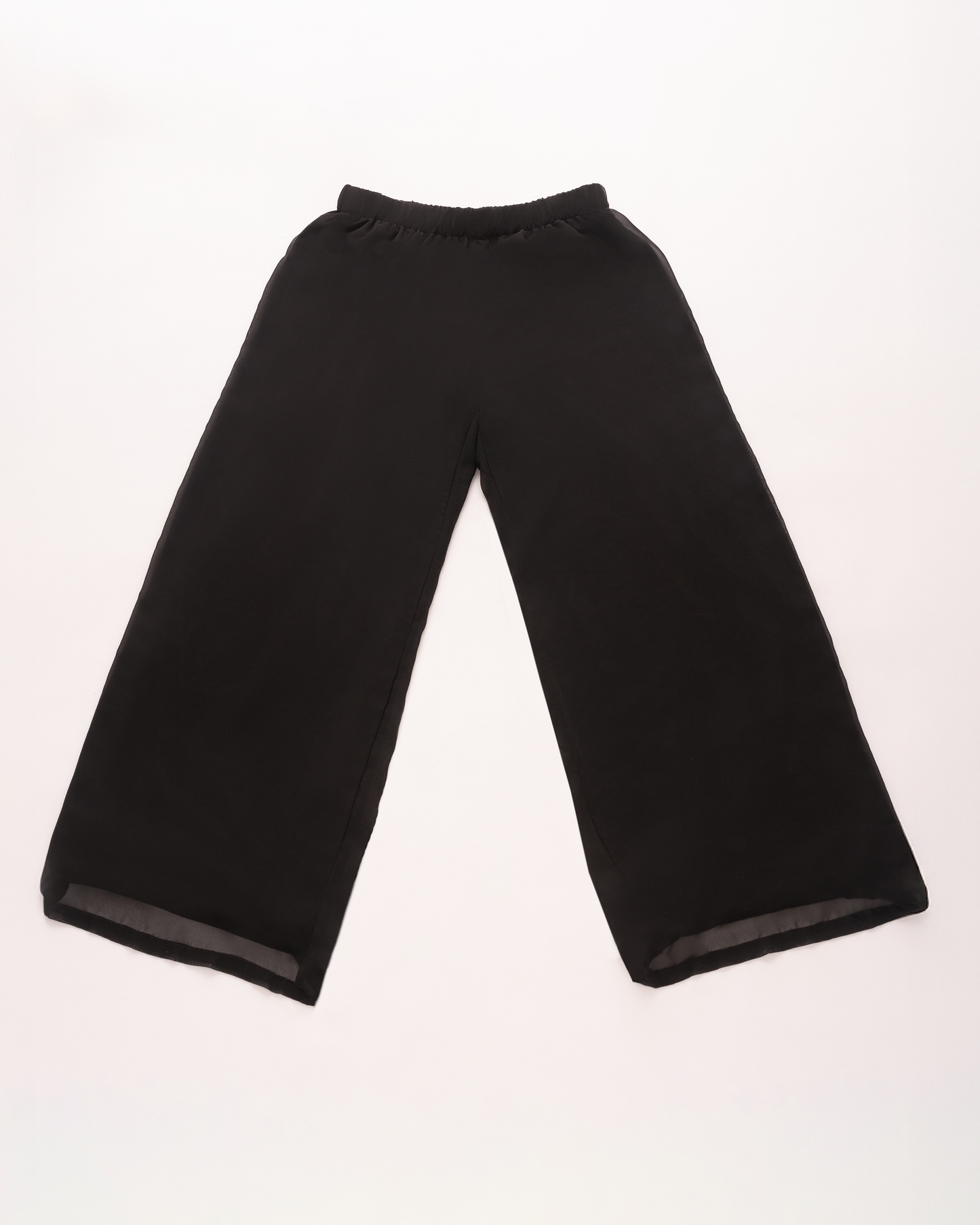 Capa plana de pantalons negres de cama ample amb una silueta relaxada, una cintura elàstica suau i una capa de gasa transparent visible a la vora, amb un fons clar.