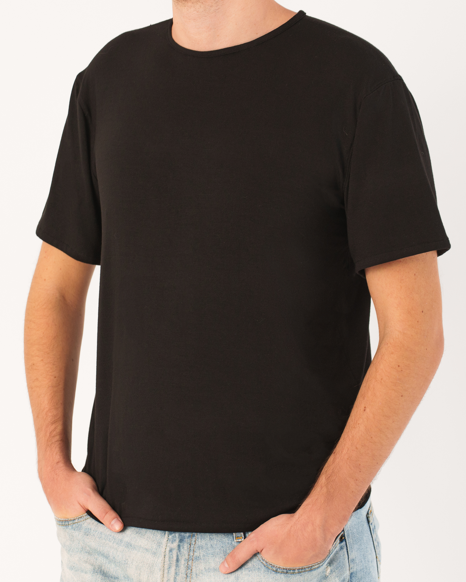 Man wearing a black short-sleeve T-shirt with a relaxed, minimalist fit, standing casually with his hands in the pockets of light blue jeans against a light gray background.