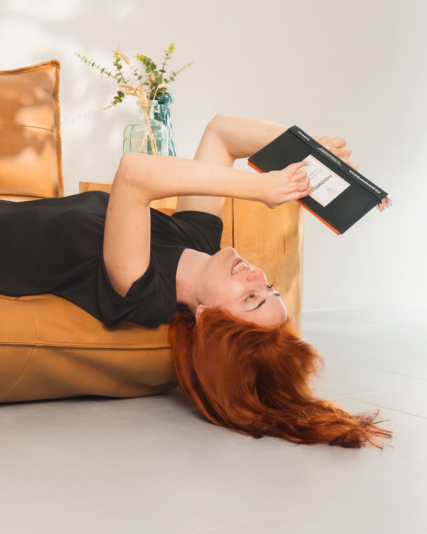 Dona amb llargs cabells vermells amb un vestit negre fluix, estirada cap per avall a la vora d'un sofà color bronzejat mentre llegeix un llibre, somrient en una postura relaxada i juganera en un interior suaument il·luminat.