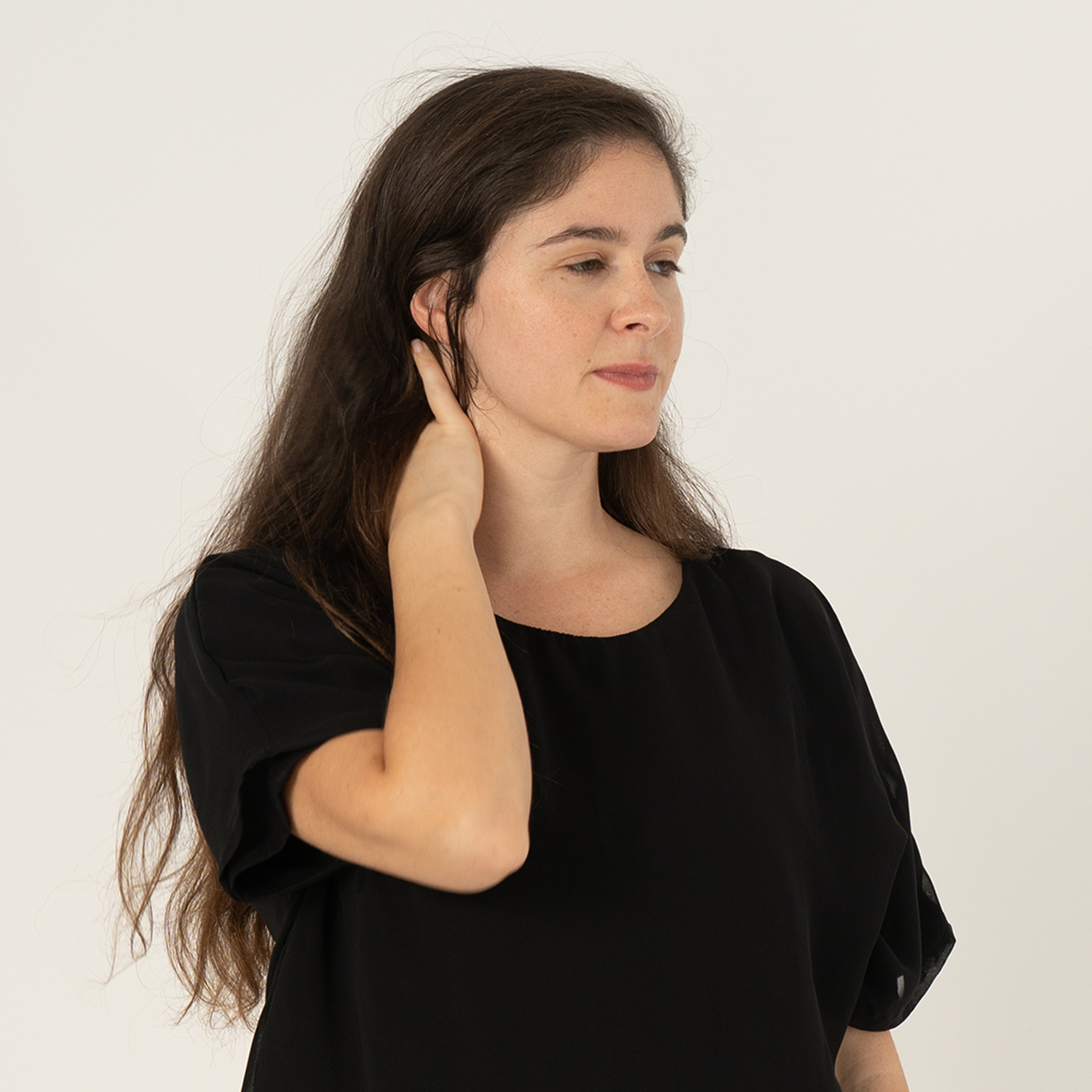 Woman with long brown hair wearing a loose black top, gently touching the back of her neck while standing against a light background.