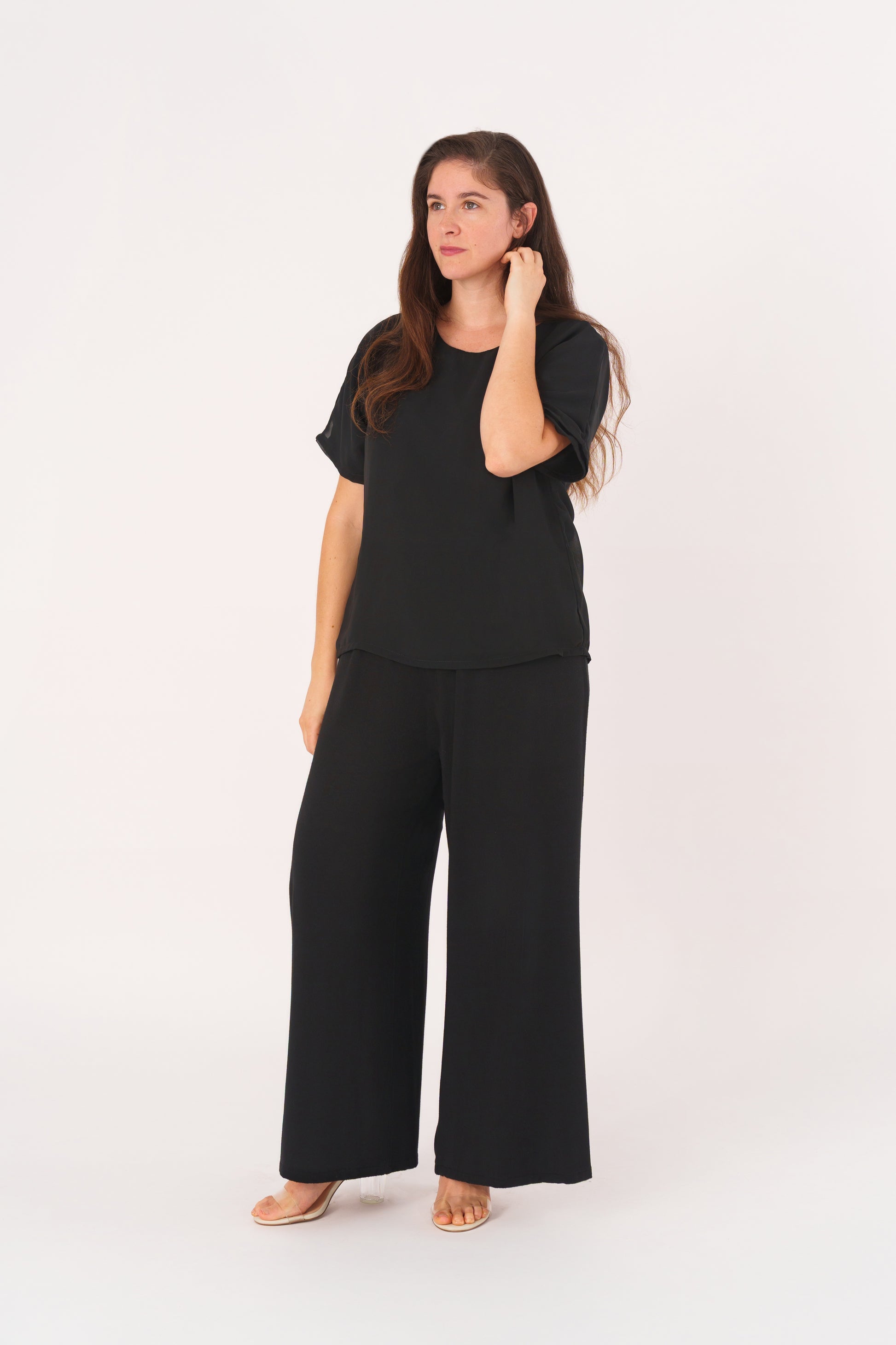 Woman with long brown hair wearing a loose black two-piece outfit with wide-leg pants and a double-layer short-sleeve top featuring a sheer chiffon outer layer, gently touching her hair while standing in a relaxed, comfortable pose against a light gray background.