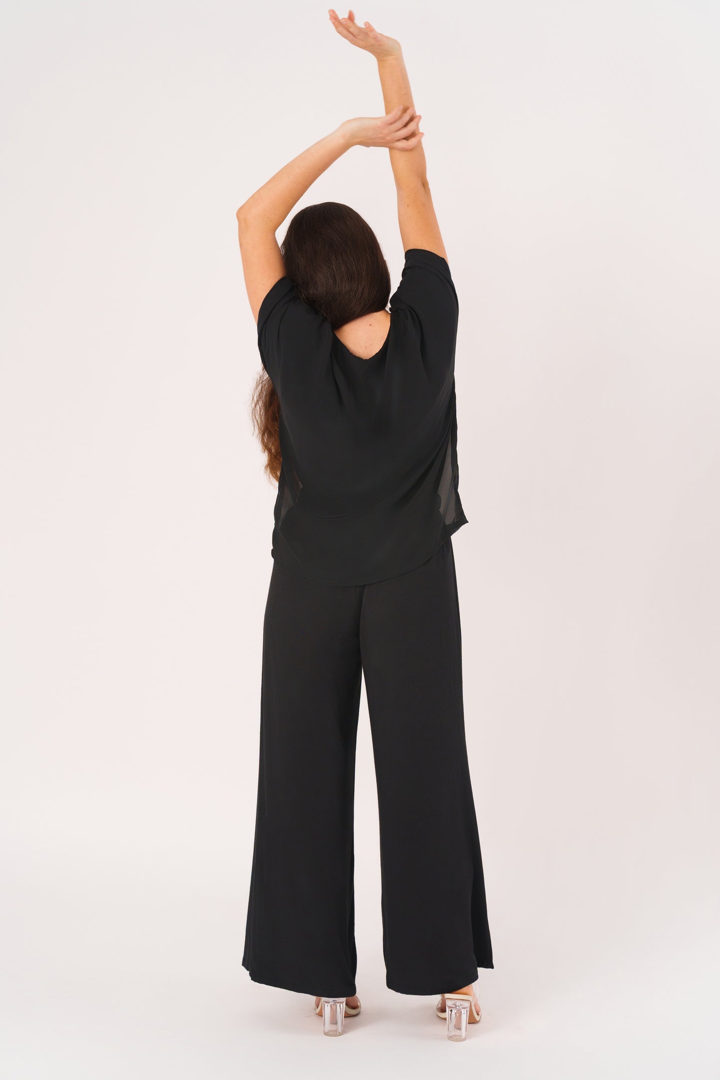 Back view of a woman with long brown hair wearing a loose black outfit with wide-leg pants and a double-layer top featuring a sheer chiffon outer layer, standing with arms raised against a light gray background.
