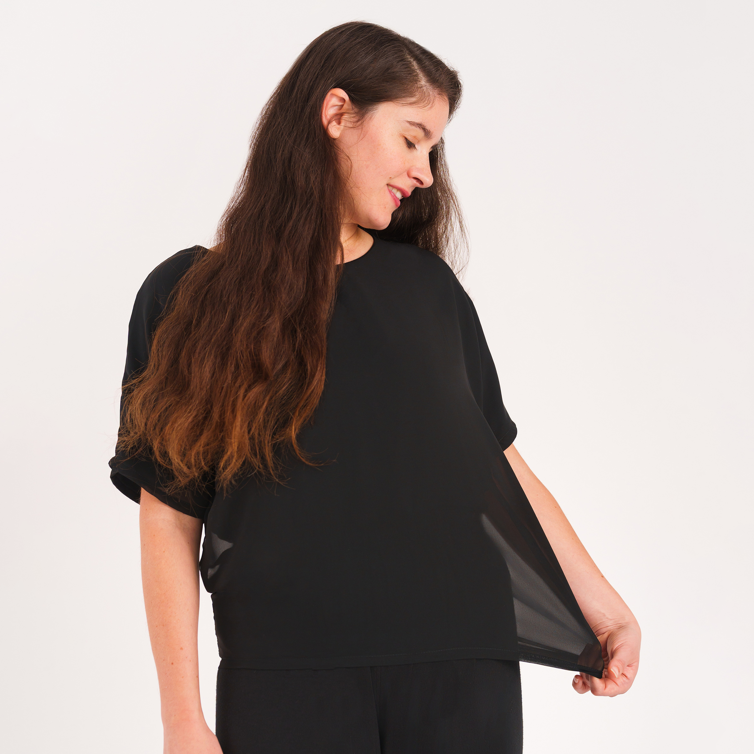 Woman with long brown hair wearing a loose black short-sleeve top with a double-layer design and sheer chiffon outer layer over matching black pants, gently pulling the lightweight fabric while looking down with a soft smile against a light gray background.