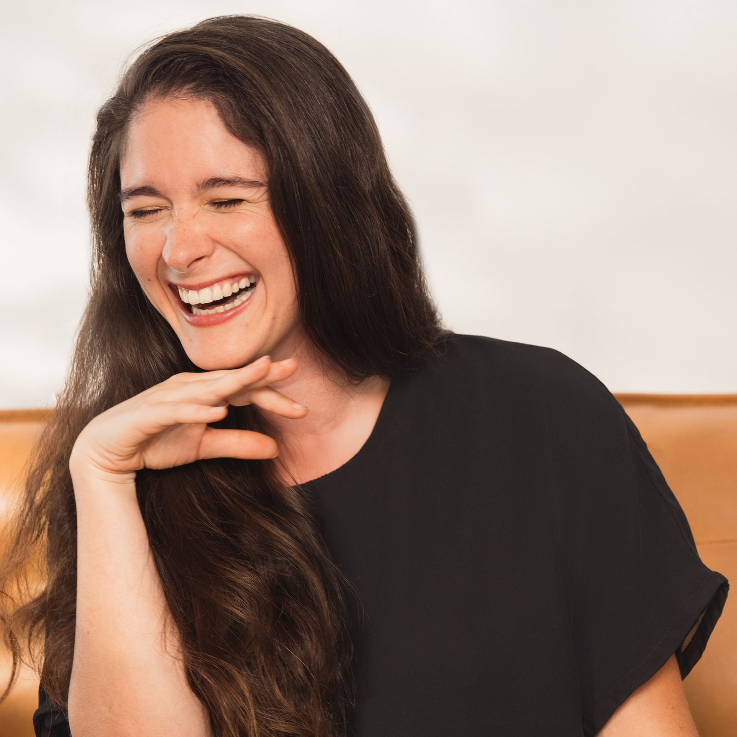 Close-up of a smiling woman with long brown hair wearing a black top, laughing with her eyes closed and her hand resting gently under her chin against a softly lit background.